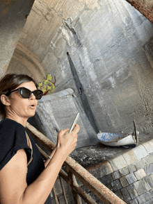 a woman wearing sunglasses looks at her phone in front of a concrete wall