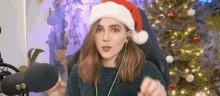 a woman wearing a santa hat and headphones is sitting in front of a microphone in front of a christmas tree .