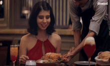 a woman in a red dress sits at a table while a waiter serves her food with dinegut written on the bottom right