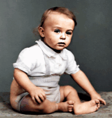 a baby in a white shirt and suspenders sits on a blanket