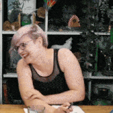 a woman wearing glasses and a black tank top laughs in front of a shelf with a book titled tranquil