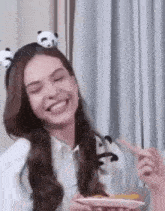 a woman wearing a panda headband is smiling while holding a plate of food and a fork .