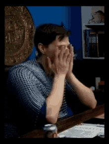 a man covering his face with his hands while sitting at a desk with a can of beer in front of him