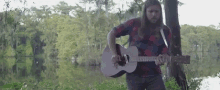 a man with long hair and a beard is playing a guitar in front of a body of water .