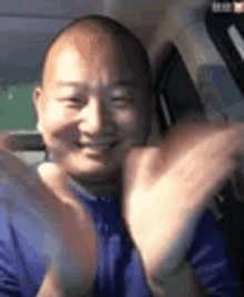 a bald man in a blue shirt is sitting in a car clapping his hands .