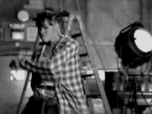 a black and white photo of a man in a plaid shirt dancing in front of a ladder .