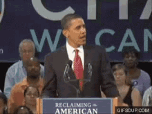 a man in a suit and tie stands at a podium with a sign that says " reclaiming american "