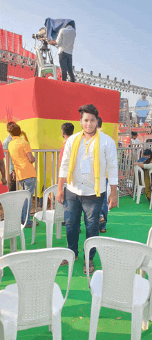 a man with a yellow scarf around his neck stands in front of empty white chairs