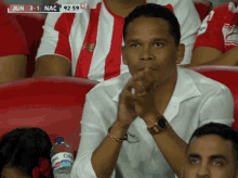 a man in a red and white striped shirt sits in a stadium watching a game between nac and jun