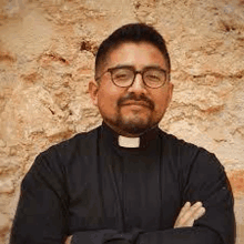 a priest with a beard and glasses is standing in front of a stone wall .