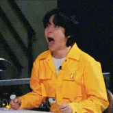 a man in a yellow jacket is sitting at a table with his mouth open and a microphone in his hand .
