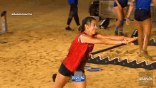 a woman in a red shirt is doing squats on a sandy beach with the word exatlon on the bottom