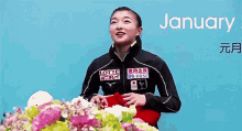 a woman in a black jacket is standing in front of a table with flowers and a blue background .