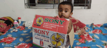 a baby is sitting on a bed next to a sony brand fan