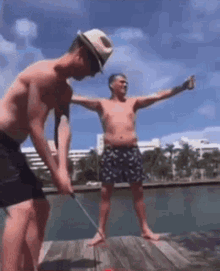 a shirtless man is swinging a golf club while another man stands on a dock with his arms outstretched .