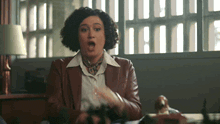 a woman in a brown jacket and white shirt is sitting at a desk with her mouth open