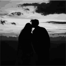 a man and woman are kissing in front of a cloudy sky in a black and white photo .