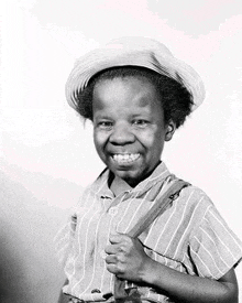 a black and white photo of a young boy wearing a hat and suspenders and smiling .