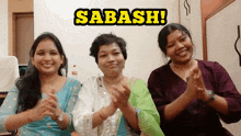 three women are clapping in front of a sign that says sabash