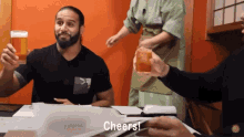 a man sitting at a table holding a glass of beer and another man holding a glass of whiskey says cheers