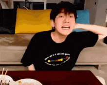 a young man is sitting at a table with a plate of food and a couch in the background .