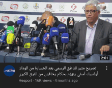 a man in a suit stands in front of a bunch of microphones and a sign that says fitco on it