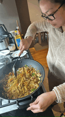 a woman is stirring noodles in a frying pan with a yellow bottle in the background