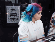 a woman with blue and pink hair sits at a table in front of a sign that says black lives matter