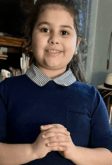 a little girl wearing a blue shirt with a striped collar is smiling with her hands folded