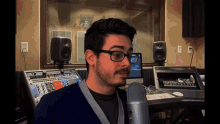 a man wearing glasses and a blue sweater stands in front of a microphone in a recording studio