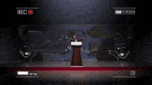a man stands at a podium giving a speech in front of a screen that says rec