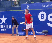 a man in a red shirt is holding a tennis racquet in front of a blue wall that says estrella damm