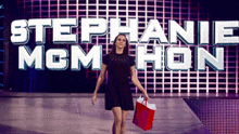 a woman in a black dress holds a red bag in front of a sign that says stephanie mcmahon