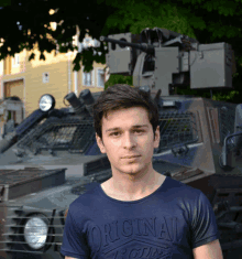 a man wearing a blue shirt that says original looks stands in front of a military vehicle