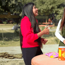 a woman in a red sweater laughs while standing next to a table with a bag of chips on it