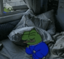 a green frog in a blue shirt is sitting on a bed next to a window .