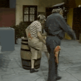 a man in a striped shirt is standing next to a barrel while another man in a uniform stands behind him .