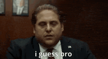 a man in a suit and tie is saying `` i guess bro '' while sitting in a chair .