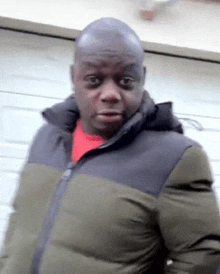 a man in a green and black jacket is standing in front of a garage door and making a funny face .