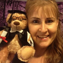 a woman smiles while holding a teddy bear wearing a graduation cap and gown