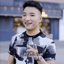 a young man in a camouflage shirt is holding a bottle of water and smiling .