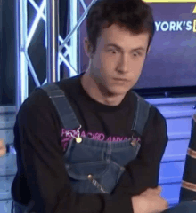 a young man wearing overalls and a black shirt is sitting with his arms crossed in front of a television .