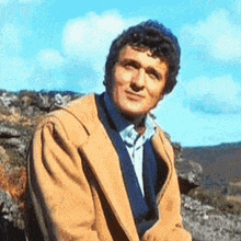 a man wearing a tan jacket and a blue shirt is sitting on a rock looking at the camera .