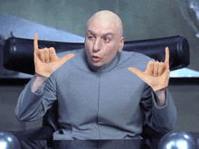 a bald man sitting in a chair making a devil horns gesture with his hands