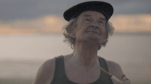 an elderly man wearing a hat and tank top holds a pipe