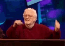 an elderly man in a red sweater is sitting in front of a purple background .