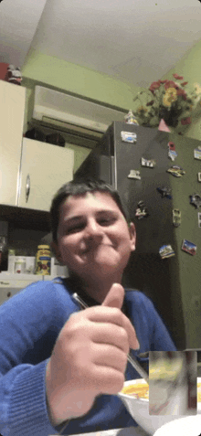 a boy in a blue sweater is giving a thumbs up while eating a bowl of cereal