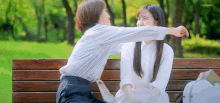 two women are sitting on a wooden bench in a park and one of them is scratching the other 's face .