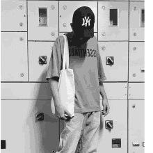 a man wearing a ny hat and a tote bag stands in front of a wall of lockers