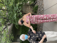 a woman in a pink floral jumpsuit and a woman wearing a blue mask shake hands